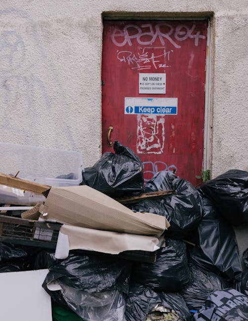 A large pile of black rubbish bags and discarded waste materials, including cardboard and plastic items, is situated directly in front of a red, weathered metal door embedded in a textured, off-white wall. The door has several graffiti tags in white spray paint and multiple stickers, one of which warns against the cost of unauthorized disposal, while another advises to keep the area clear. The trash pile extends across the lower part of the image, overlapping the bottom of the door, and suggests an instance of on-site clearance or private waste disposal rather than council collection. The environment appears to be an outdoor urban alleyway or service area, with no visible surrounding structures or greenery. The lighting is natural, evenly illuminating the scene, emphasizing the textures and colors of the debris and wall, consistent with private rubbish handling or independent rubbish removal services such as those provided by Rubbish Collection Lambeth.