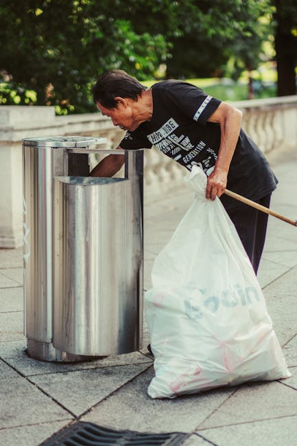 A man with dark hair and wearing a black T-shirt with white text is seen leaning over a metallic outdoor rubbish bin, actively disposing of waste. He is holding a large white garbage sack in his left hand, which appears to contain waste materials. The bin is made of stainless steel with a shiny, reflective surface, and is positioned on a paved sidewalk. In the background, there are lush green trees and a low stone wall, suggesting an outdoor urban or park setting. The scene is illuminated by natural daylight, creating a bright and clear atmosphere. This image exemplifies practices related to private waste handling and rubbish disposal services, such as those offered by Rubbish Collection Lambeth, and highlights the importance of proper waste management and on-site rubbish collection in residential or public outdoor environments.