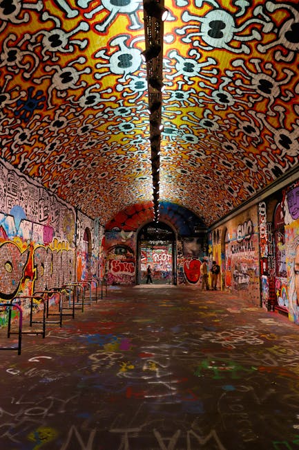 An underground tunnel with a curved ceiling covered in vibrant, intricate graffiti art featuring bold patterns, stylized skull motifs, and swirling designs in bright orange, yellow, black, and white. The tunnel's floor is also decorated with various colorful spray-painted markings and tags, creating a lively and chaotic atmosphere. On the left side, there are metal benches with a simple design positioned against the graffiti-covered wall, while the right side features additional graffiti and some silhouetted figures standing near the far end of the passage. A black pole or lighting fixture runs down the center of the ceiling, dividing the tunnel visually. The scene is illuminated with natural and artificial light sources, highlighting the detailed textures of the spray paint and the smooth, curved surface of the tunnel. Although the setting appears to be an urban or artistic space rather than a typical rubbish removal environment, it exemplifies an area often associated with alternative forms of urban art and street culture, aligning with themes of independent or private creative spaces in a city context.