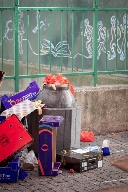 A collection of discarded rubbish positioned against a concrete base beneath a green metal fence featuring graffiti artwork. The waste includes a purple cardboard box labeled 'IRA FRUIT', a black plastic container, a red plastic crate tilted on its side, and various smaller debris such as plastic bags and torn packaging. An old ceramic pot filled with torn red fabric or paper goods is placed atop the concrete block. Surrounding the main rubbish are additional scattered items, including a red plastic bag on the cobbled pavement near the curb and some small fragments of debris. The scene suggests informal littering in an outdoor urban environment, potentially requiring scheduled rubbish collection or alternative waste disposal services. The background physical environment consists of the textured metal fence with white outline graffiti figures, indicating a public space or street front likely encountered in Lambeth, and the overall atmosphere reflects typical city street cleanup needs that professional waste removal services could address, such as those offered by Rubbish Collection Lambeth.