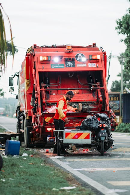 A sanitation worker in a red and yellow uniform is shown collecting waste near the rear of a large red rubbish collection truck parked beside a street. The truck features a wide, flat loading area with mechanical arms and hydraulic components visible, and its surface is slightly weathered with visible dirt and grime. The worker is bent over, handling a black plastic trash bag placed on a small, portable waste collection platform attached to the truck. Surrounding the scene, there is a paved roadside with a white line marking the edge, and some scattered debris on the ground. In the background, a few trees, electrical wires, and a blue building with signage are visible, indicating an urban environment. The lighting appears natural, suggesting the image was taken during daylight hours, and the scene reflects an on-site waste collection operation, aligning with the context of independent rubbish removal services like those offered by Rubbish Collection Lambeth.