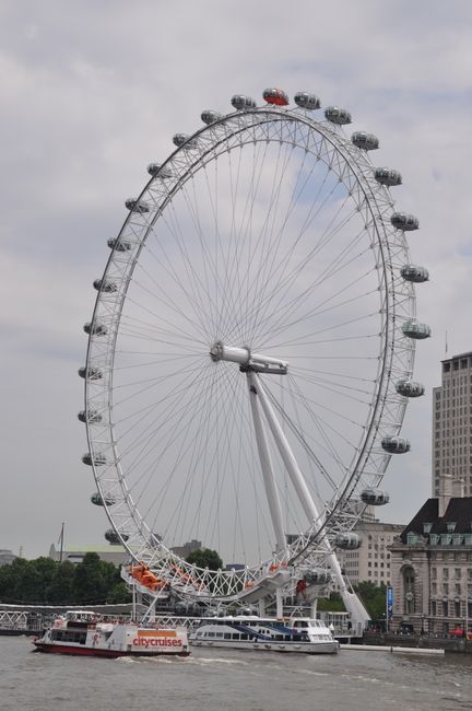 A large Ferris wheel with a metal frame and multiple enclosed passenger cabins, some of which are red, gray, and clear, situated near a river. The Ferris wheel's structure includes numerous spokes connecting the central hub to the outer rim, supported by a prominent diagonal support arm. The cabin gondolas are evenly spaced around the circular track, with some cabins visible on the right side of the wheel. In the background, there are several mid-rise buildings with mixed architectural styles, including a historic-looking stone structure and a modern high-rise, under a cloudy sky. At the base of the Ferris wheel, a white river cruise boat labeled 'city cruises' is docked, indicating a waterfront area that might occasionally be used for private or alternative forms of sightseeing or leisure activities. The scene appears to be in an urban setting, with the Ferris wheel serving as a notable landmark near the water, providing a contrast between amusement infrastructure and cityscape environments, which might be relevant to discussions about different types of waste management or disposal options in busy tourist areas.