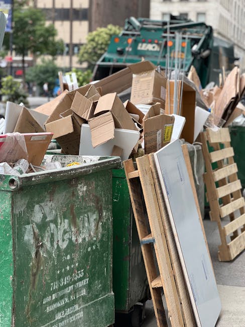 A large, open green waste skip filled with various discarded cardboard boxes, some flattened and others in their original form, along with a few white foam packaging pieces and smaller paper debris. The boxes are in different shades of brown, with some showing printing or labels. Leaning against the side of the skip are wooden pallets, typically used for transporting goods, positioned vertically and extending slightly above the rim. In the background, a rear loading waste collection vehicle with a teal and black finish is partially visible, parked on an urban street lined with multi-storey buildings. The scene takes place outdoors during daylight hours, with natural lighting illuminating the cluttered waste scene, characteristic of private or independent rubbish clearance services often carried out by companies like Rubbish Collection Lambeth. Surrounding the area, there are trees and a few parked vehicles, indicating a busy city environment. The image captures a typical scene of refuse awaiting collection, reflecting the operational activities related to rubbish and waste management in the city center.
