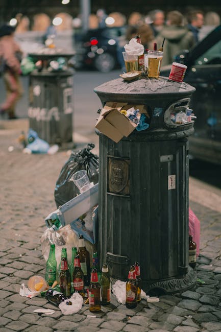 A black cylindrical metal rubbish bin with a weathered surface sits on a cobbled pavement in an urban setting. The lid is closed, and the bin is surrounded by various types of discarded waste, including crumpled paper, empty glass bottles, and plastic wrappers, some spilling onto the ground. Several beverage bottles, primarily green and brown glass, are placed around the base of the bin, with some leaning against it. On top of the bin's lid, there are multiple paper cups and a snack container, some with remaining drink residue and food remnants. Adjacent to the bin, a black plastic trash bag is tied and partially open, with additional waste visible nearby. In the background, there is a blurred view of parked cars, a grey waste collection bin, and unfocused pedestrians in an outdoor city environment, indicating a busy street scene that might require informal waste handling or communal disposal. The lighting suggests late afternoon or early evening, with natural daylight illuminating the scene, consistent with urban waste accumulation in public areas.