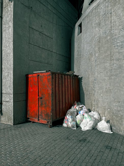A large, rectangular metal skip with a weathered red paint finish, situated against a dark green wall and a plain, light grey concrete wall in an outdoor urban setting. The skip appears to be used for waste disposal and is positioned on a paved surface with small, interlocking grey bricks. Several plastic trash bags filled with miscellaneous refuse are placed on the ground beside the skip, some slightly crumpled and varying in transparency. The skip's metal surface shows signs of rust and chipped paint, indicating frequent use. The surrounding environment suggests an alleyway or service area, with minimal natural light visible. This scene exemplifies a typical private waste collection scenario, where rubbish is temporarily stored before being collected by a waste management company such as Rubbish Collection Lambeth. The background features a dark green wall on the left side and a plain concrete wall behind the skip, highlighting a utilitarian, functional space designated for rubbish storage and disposal.
