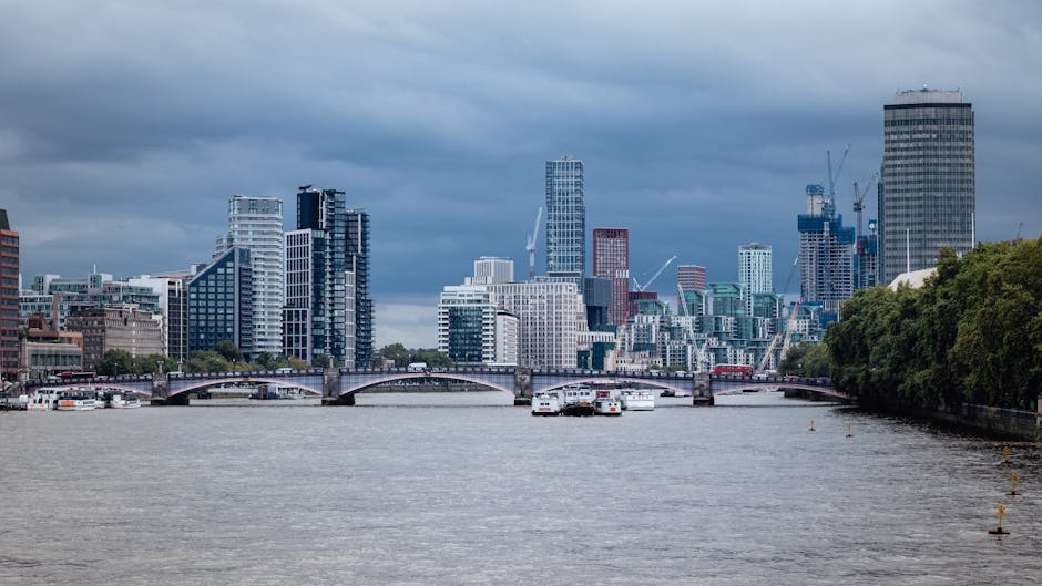 A panoramic daytime view of a cityscape along a river, featuring a variety of modern high-rise buildings with glass facades and steel structures. Some skyscrapers are under construction with visible cranes on top. The river in the foreground is calm, with several white boats moored along the bank. A stone bridge with multiple arches spans the river, connecting two sections of the city. The right side of the image shows a lush green tree-lined bankside, contrasting with the urban environment. Overhead, dark storm clouds cast a subdued lighting across the scene. This image, associated with rubbish removal services, highlights the importance of maintaining urban cleanliness and could imply the need for proper waste collection in busy city environments, whether through council services or private waste handling solutions like those provided by Rubbish Collection Lambeth.
