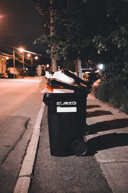 A black 240-litre wheelie bin with the label 'St. John's' on the front, positioned on a paved sidewalk next to a curb in an urban residential area at night. The bin is filled with various types of rubbish, including flattened cardboard boxes, plastic bags, and miscellaneous waste materials, some of which are protruding from the open lid. The background features dimly lit streetlights, a tree with dark leafy branches casting shadows, and a row of residential houses and parked cars along the street. The scene suggests a typical outdoor setting for private or independent rubbish disposal, with the bin awaiting collection. The environment is calm, with artificial lighting providing contrast against the dark sky, emphasizing the bin and surrounding pavement in a subtle context of waste management and rubbish removal services.