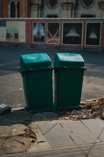 Two green plastic rubbish bins with domed lids and black handles are positioned side by side on a sidewalk near the edge of a paved area, with a slightly raised curb. The bins are made of durable, textured plastic and are slightly weathered, showing minor scuffs and dirt on their surfaces. They stand close to a small pile of dried leaves and organic debris, indicating recent placement or collection. Behind the bins, a tarmac road is visible, with a brick and stone building facades featuring decorative windows and architectural details in the background. The scene is lit by natural daylight, with shadows cast on the pavement, suggesting a clear day. The environment appears to be an urban or suburban street, and the presence of the bins alongside a pavement and road indicates normal waste collection activity, possibly an alternative to communal waste disposal methods, aligned with private waste handling services like those provided by Rubbish Collection Lambeth.