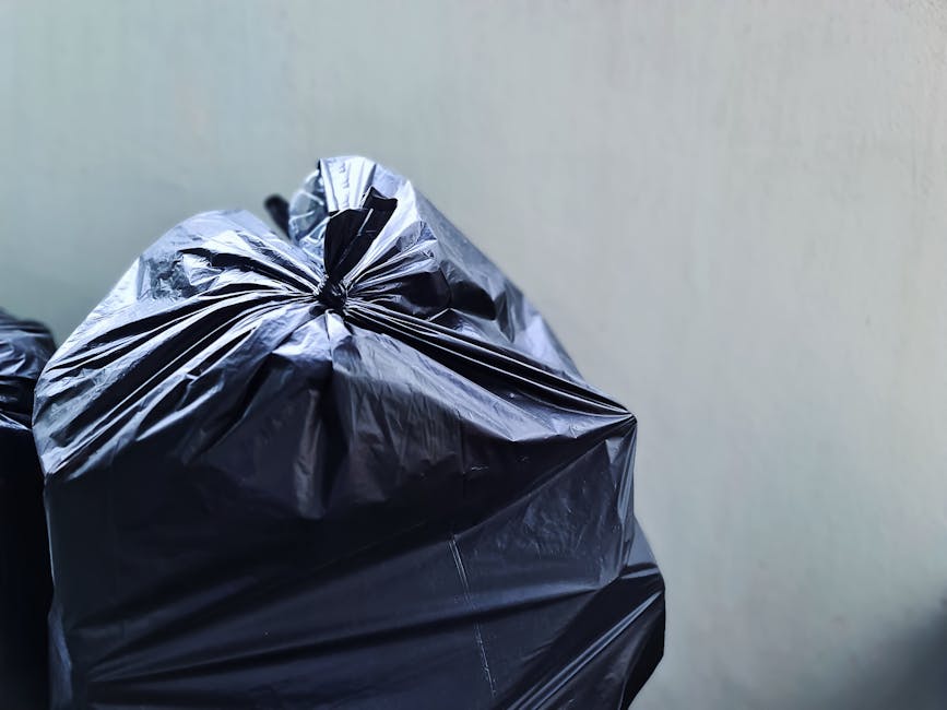 A black 240-litre wheelie bin with the label 'St. John's' on the front, positioned on a paved sidewalk next to a curb in an urban residential area at night. The bin is filled with various types of rubbish, including flattened cardboard boxes, plastic bags, and miscellaneous waste materials, some of which are protruding from the open lid. The background features dimly lit streetlights, a tree with dark leafy branches casting shadows, and a row of residential houses and parked cars along the street. The scene suggests a typical outdoor setting for private or independent rubbish disposal, with the bin awaiting collection. The environment is calm, with artificial lighting providing contrast against the dark sky, emphasizing the bin and surrounding pavement in a subtle context of waste management and rubbish removal services.