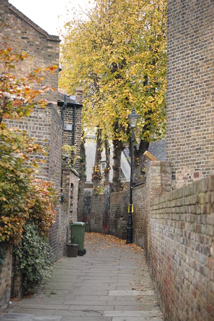 A narrow brick-paved alleyway in an urban residential area during autumn, flanked by high brick walls on both sides. On the left, there is a small shrub with orange and green leaves, and a green wheelie bin positioned near the wall. At the end of the alley, a large deciduous tree with yellow and orange foliage extends its branches overhead, partially obscuring the view beyond. A black street lamp with a classic design is mounted on a pole along the right wall, which is topped by a brick wall of a building structure. The environment appears quiet and well-maintained, with fallen leaves scattered on the pavement. The scene exemplifies an independent alleyway, often requiring clearance or waste removal in residential management, relevant to non-local authority waste handling by services such as Rubbish Collection Lambeth, especially when considering household waste or garden clearance in the Lambeth area.