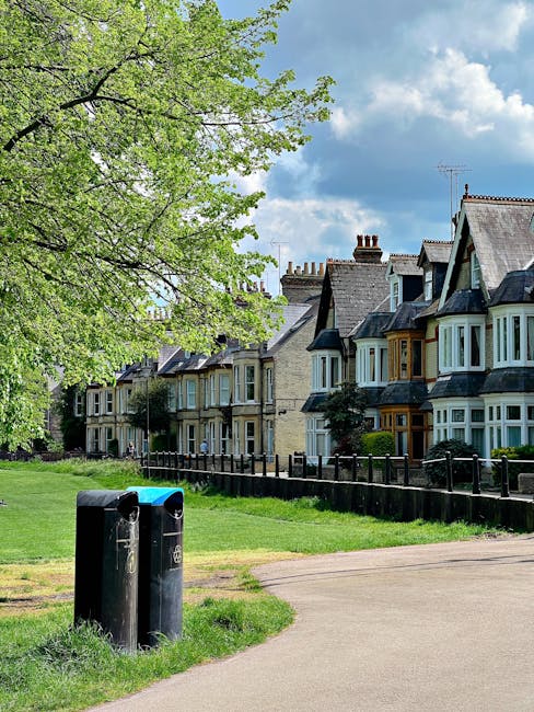 A row of traditional terraced houses with bay windows, pitched roofs, and chimneys, situated along a quiet residential street. In the foreground, two black rubbish bins with rounded lids and small openings are positioned on a gravel pathway next to a grassy verge. A large deciduous tree with fresh green foliage extends its branches over the scene on the left side, casting partial shade. The background features a partly cloudy sky with patches of blue visible through white clouds, and the houses are bordered by a low wooden fence. The environment appears calm, typical of suburban neighbourhoods, suggesting that the scene relates to local rubbish collection or private waste disposal services often required for house clearance or on-site rubbish management, with Rubbish Collection Lambeth providing relevant waste removal solutions in areas like this. The overall scene emphasizes the importance of proper waste management in residential areas, consistent with private rubbish services offering alternative disposal options to municipal collection.