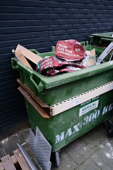 A large green wheeled rubbish bin positioned on a paved surface beside a dark brick wall, filled with red and white bags of garden or building waste marked with text such as 'FK200 FLEXY PLUS.' The same green bin is stacked on top of a white communal recycling container with green markings and a website address, indicating an independent waste collection service. The scene suggests an area designated for private or alternative rubbish removal, with additional equipment, such as a metal ramp or platform, partially visible at the bottom of the image. The environment appears to be an outdoor space, possibly a back alley or side yard, with natural lighting casting soft shadows. The surroundings, including the pavement and wall, indicate a typical urban setting where waste disposal or collection arrangements might be made outside residential or commercial premises, reflecting practical waste management procedures.