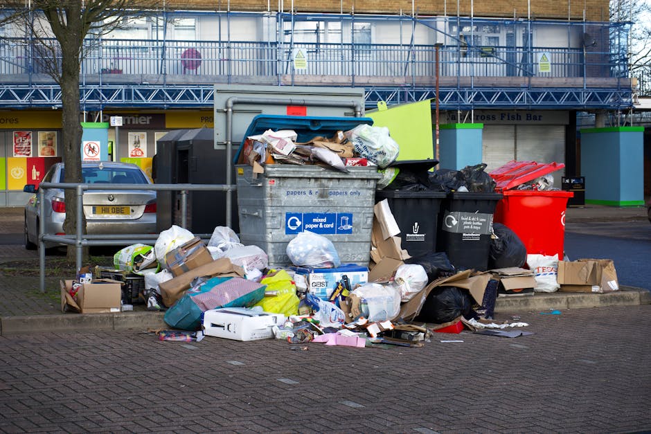 A collection of overflowing rubbish bins and bags is positioned on a paved sidewalk in front of a commercial building. The waste containers include a large grey mixed paper and cardboard bin, a black bin for general waste, and a bright red bin. Around these bins, there are numerous black and white plastic rubbish bags, some tied tightly and others torn open, spilling their contents onto the pavement. Cardboard boxes, paper, plastic packaging, and miscellaneous household items are scattered across the ground, indicating an uncollected or poorly managed waste situation. Behind the waste area, a parking space with a silver car is visible, and a low metal railing partially surrounds the waste collection point. In the background, the building features a storefront with signage and large windows, all framed by a blue metal scaffolding structure. The scene suggests an example of private or independent waste collection, aligning with services that handle rubbish removal outside of municipal systems, consistent with the context of alternative waste handling services.
