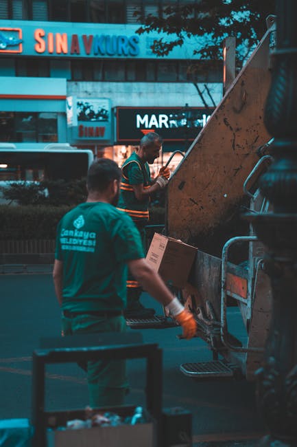 A large green wheeled rubbish bin positioned on a paved surface beside a dark brick wall, filled with red and white bags of garden or building waste marked with text such as 'FK200 FLEXY PLUS.' The same green bin is stacked on top of a white communal recycling container with green markings and a website address, indicating an independent waste collection service. The scene suggests an area designated for private or alternative rubbish removal, with additional equipment, such as a metal ramp or platform, partially visible at the bottom of the image. The environment appears to be an outdoor space, possibly a back alley or side yard, with natural lighting casting soft shadows. The surroundings, including the pavement and wall, indicate a typical urban setting where waste disposal or collection arrangements might be made outside residential or commercial premises, reflecting practical waste management procedures.