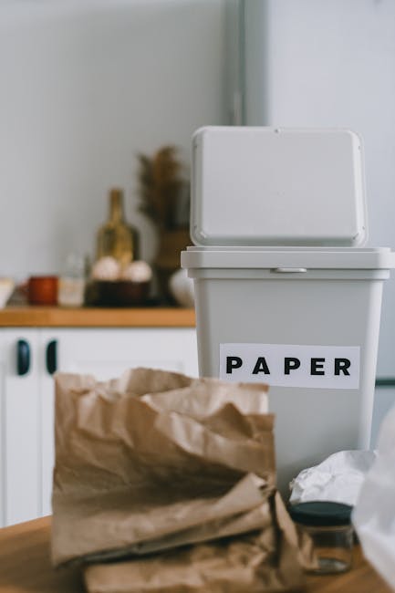 A white plastic waste bin with an open lid, positioned on a countertop in a kitchen environment, features a label with the word 'PAPER' printed in bold black letters on a white background. The bin is situated in front of a slightly blurred background that includes a light-colored wall, a wooden surface, and various kitchen items such as jars, containers, and utensils. In the foreground, there is a crumpled brown paper bag and some loose crumpled paper and a small black object, possibly a lid or cap, resting nearby. The scene is illuminated by natural or soft artificial light, highlighting the textures of the paper, the smooth surface of the bin, and the environment's neutral tones. This setup reflects an aspect of domestic waste sorting or an alternative approach to rubbish collection through private or on-site waste handling, common within home recycling practices supported by companies like Rubbish Collection Lambeth.